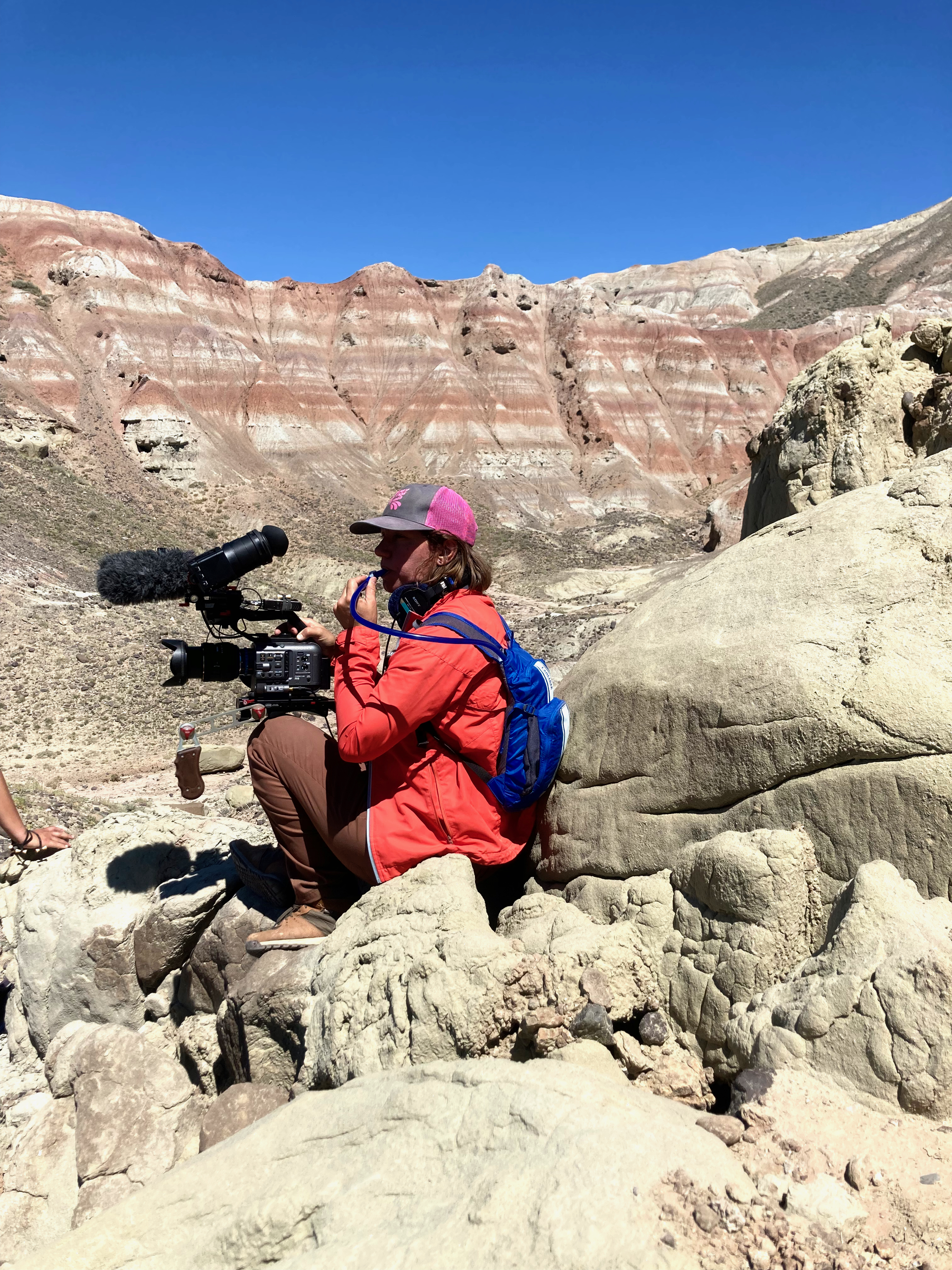 Woman in orange raincoat and pink cap holds a camera rig while drinking from straw connected to blue backpack. She sits on a boulder with a red striped cliff face behind her.