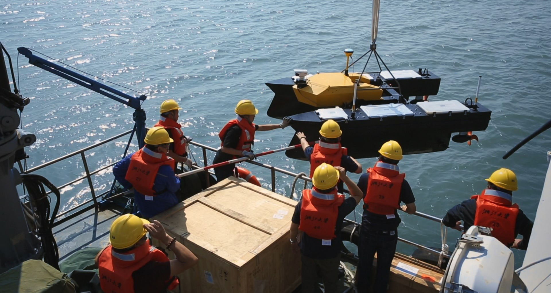 Eight men in orange vests and yellow hard hats on ship raise two containers out of the ocean