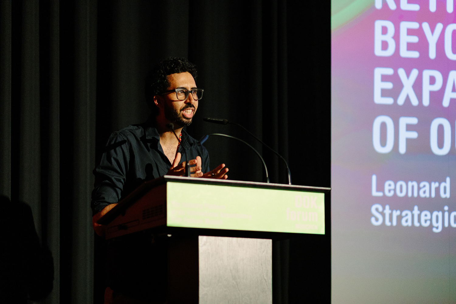 A man in a black shirt with black glasses and short dark hair and beard delivers a speech at a podium. Behind him is a black curtain and the lefthand side of a pink and green digital display showing text which cannot be read