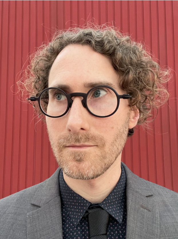 A light-skinned man wearing dark glasses is looking to the left with a timid smile. He has dark blond, curly hair. He is dressed in a gray suit and a black tie, with a red background behind him.