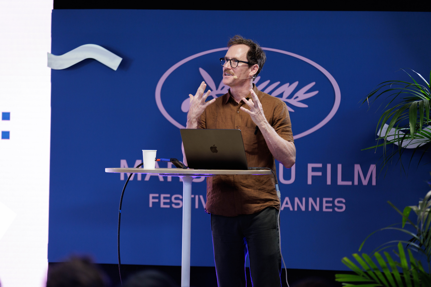 White middle-aged man with brown hair, glasses and a neat mustache wears a brown shirt against a blue backdrop at Cannes's Marche du Film standing in front of a laptop mid-performance