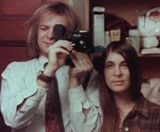 An adult male with blonde hair to the shoulders is holding a small camera, next to an adult woman with long brown hair