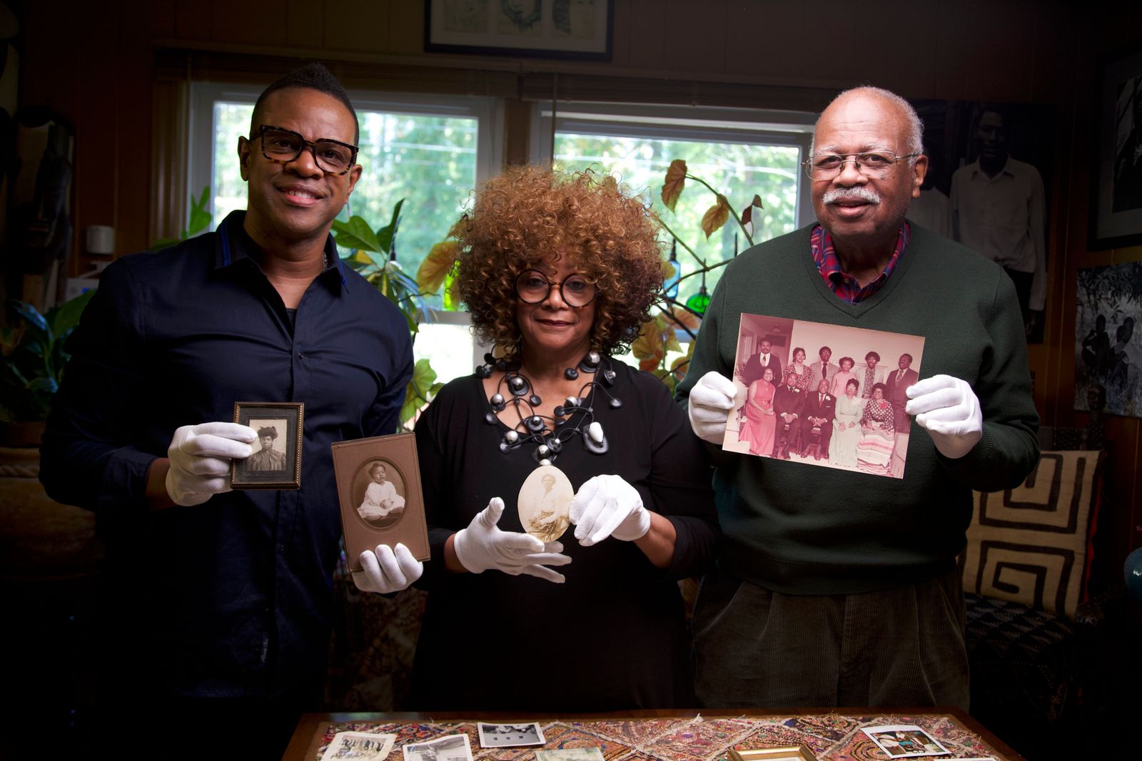 'Family Pictures USA'  artist Thomas Allen Harris, left, a Black man with glasses and a blue button-down shirt, displays family photographs belonging to North Carolina Poet Laureate Jaki Shelton Green, a Black woman with copper-colored curly hair. round glasses and a black dress, and  her cousin, Sterling Michael Holt., a Black man with white hair and white mustache, glasses, waring a green sweater and holding a family photograph.   Harris' 'Family Pictures USA' project co-creates a living and growing family picture album of America by traveling across the country and inviting community members to share images and stories from their personal family archives.  Photo courtesy of Thomas Allen Harris. 
