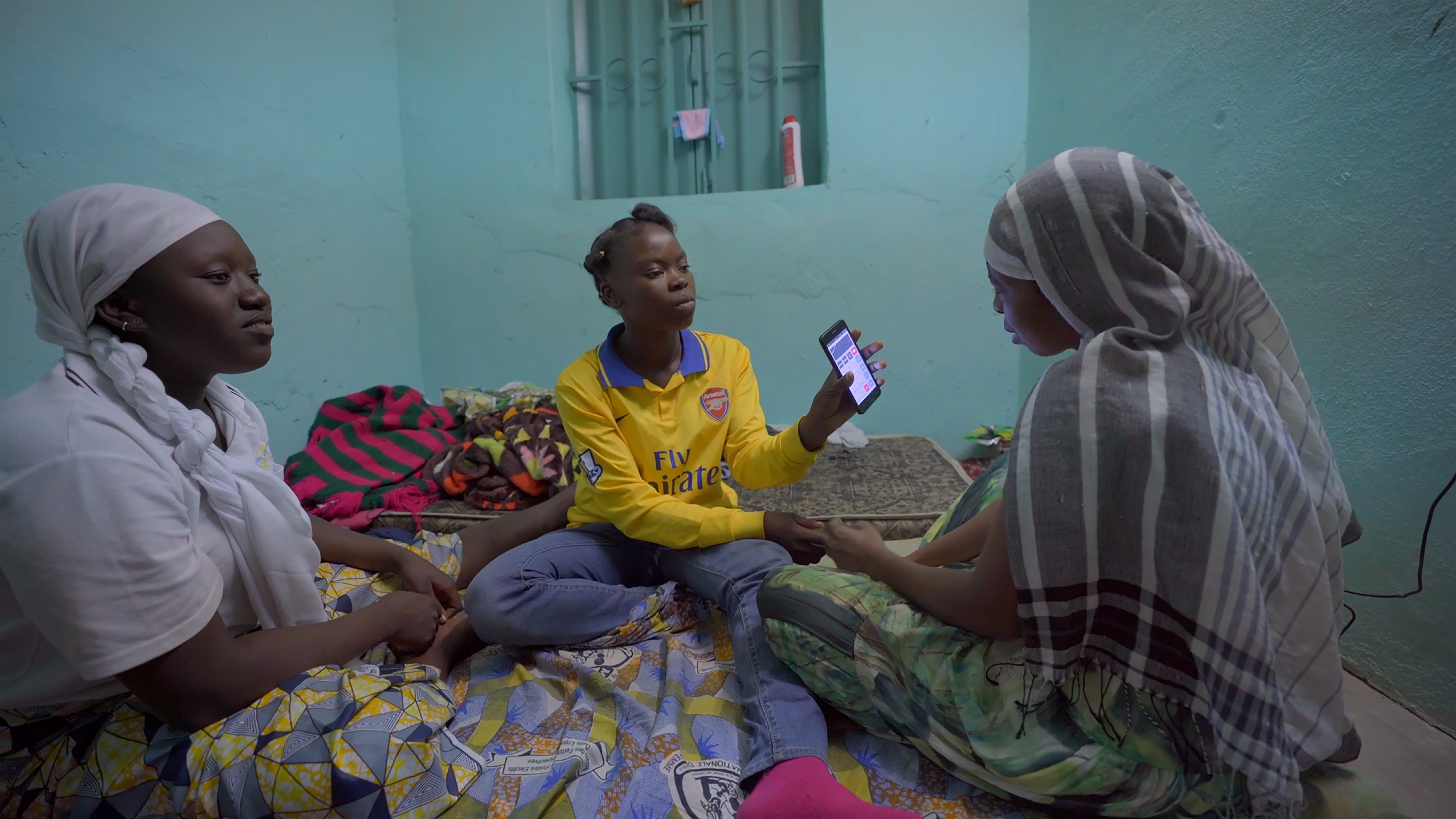 Three African girls sit in a room against a turquoise background as one shows her phone. From Ousmane Samassékou’s 'The Last Shelter.' Photo courtesy of STEPS. 