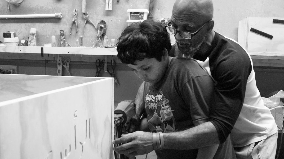 :A  bald Black man with glasses teaches a young boy with black hair how to engrave a casket with a drill.