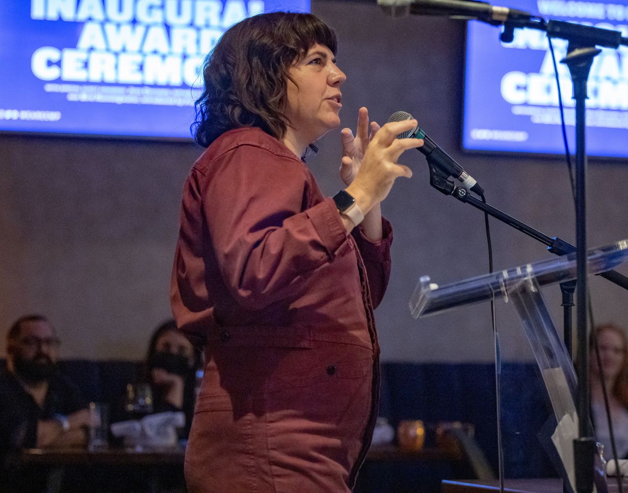 Jessica Devaney is a white woman with shoulder-length brown hair. She is wearing a rust jumpsuit and speaking into the mic at Doc 10's awards ceremony. Photo by Barry Brecheisen. 