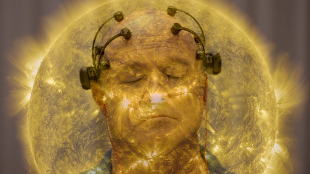 A man, who is particiapting in a neuroscience focus group, has his eyes closed, and is wearing a device on his head comprised of four neurosensors attached to the top of his head and twolarger sensors above each ear. He in enveloped in a golden aura that resembles the sun.