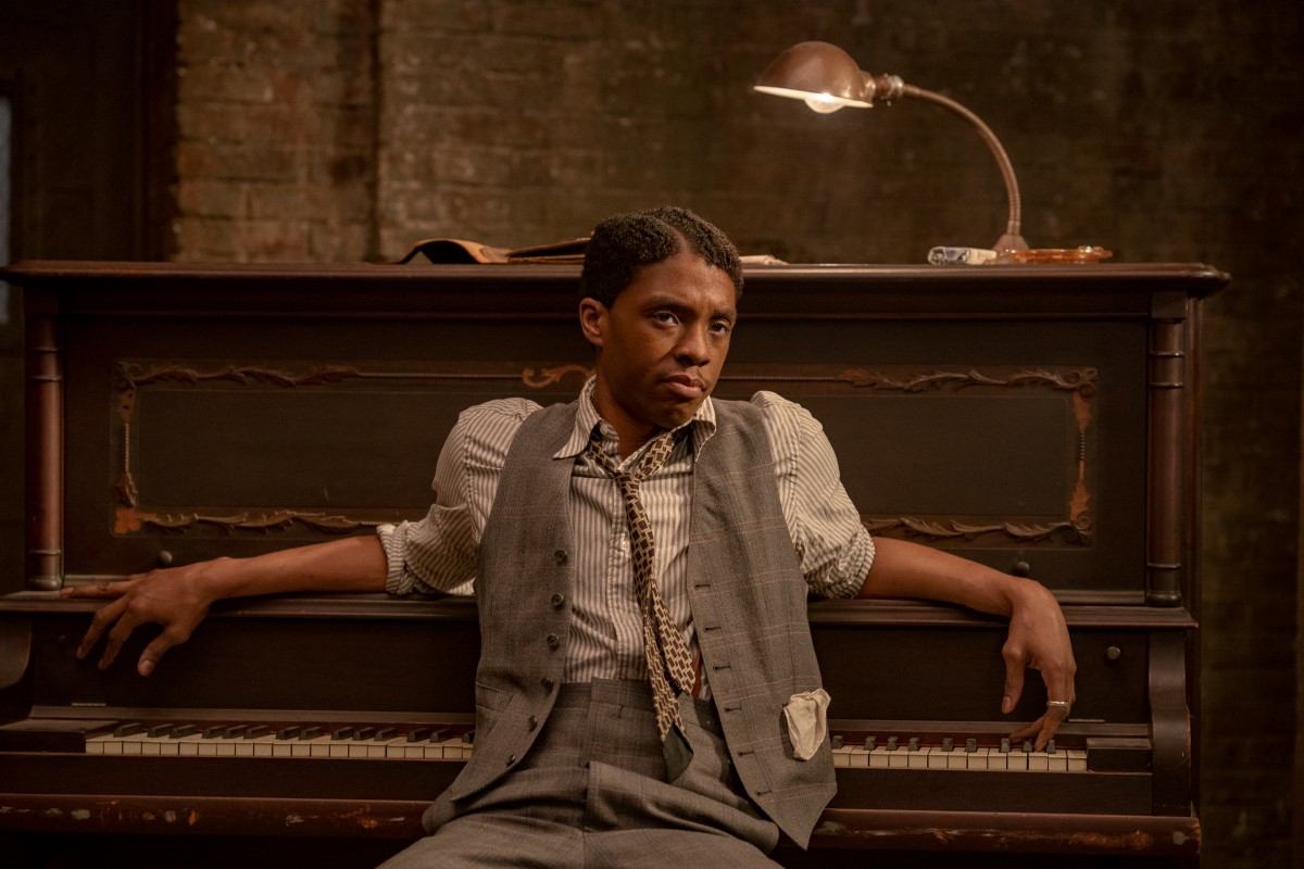 Actor Chadwick Boseman, a Black man wearing a striped shirt, patterned tie, grey vest and pants, sits at a piano, his back to the keyboard