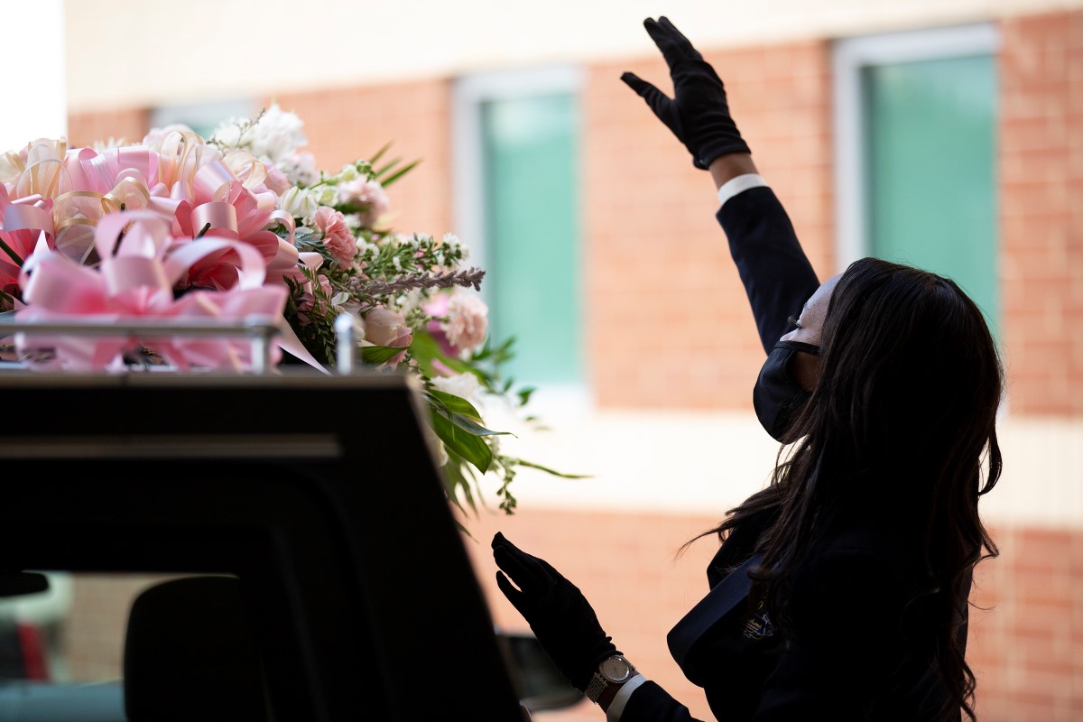 The photo depicts a Black woman standing over an open casket, her right arm raised in praise; she wears a protective face mask, as well as a dark suit.