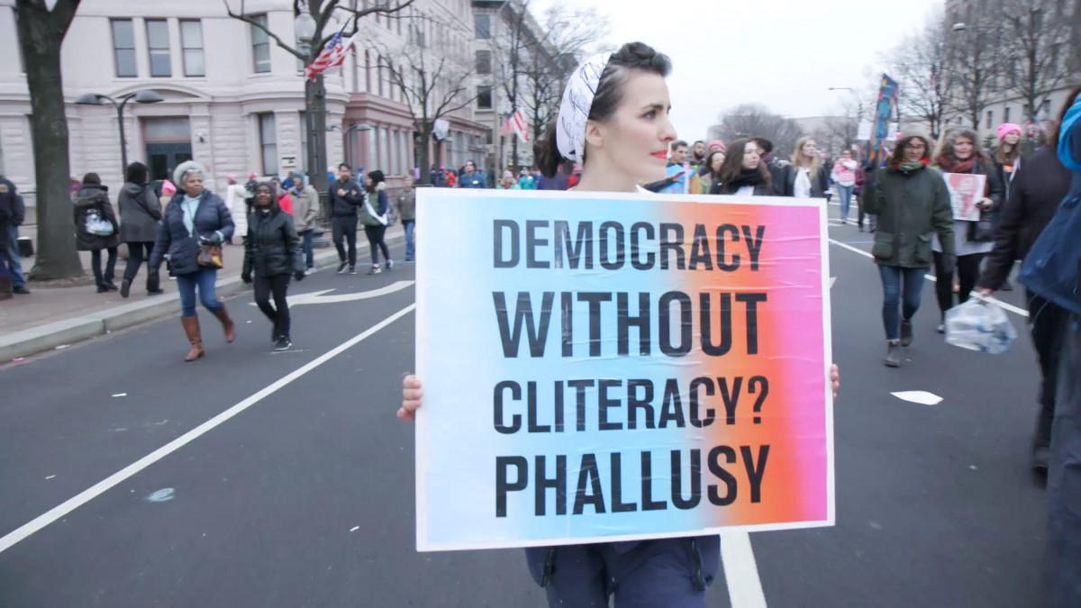 A woman is particpating in a demonstration on a city street; she carries a sign that reads "Democracy without cliteracy? Phallusy."