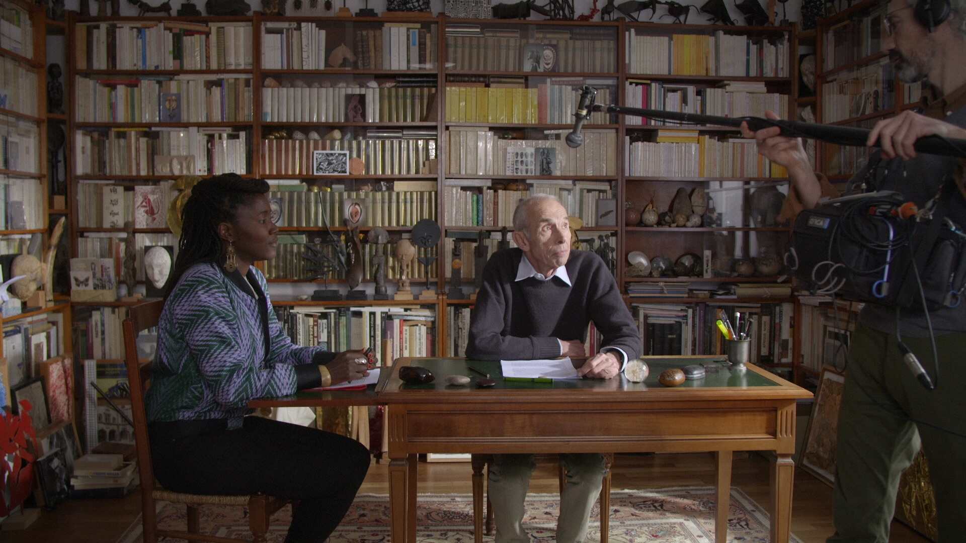 Alice Diop is a Senegalese-French woman wearing a multicolored jacket. She is sitting in a library, speaking to Pierre Bergounioux, an older white French male writer. They're both being filmed. From Alice Diop's 'Nous.' Photo by Sarah Blum. Courtesy of Mubi.