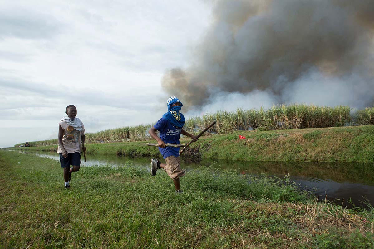 Two Black teenage boys run across a green field holding sticks. Smoke bellows behind them. Still from Patrick Bresnan's 'The Rabbit Hunt'. Courtesy of Argo