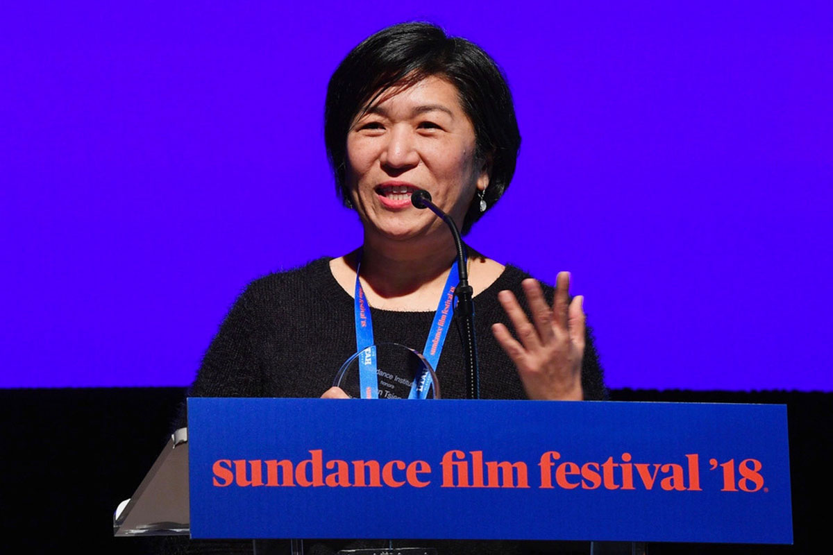 Jean Tsien is a female Asian American filmmaker seen here receiving the Art of Editing Mentorship Award at Sundance, 2018. She is speaking into a mic and wearing a black sweater. Photo courtesy of Jean Tsien.