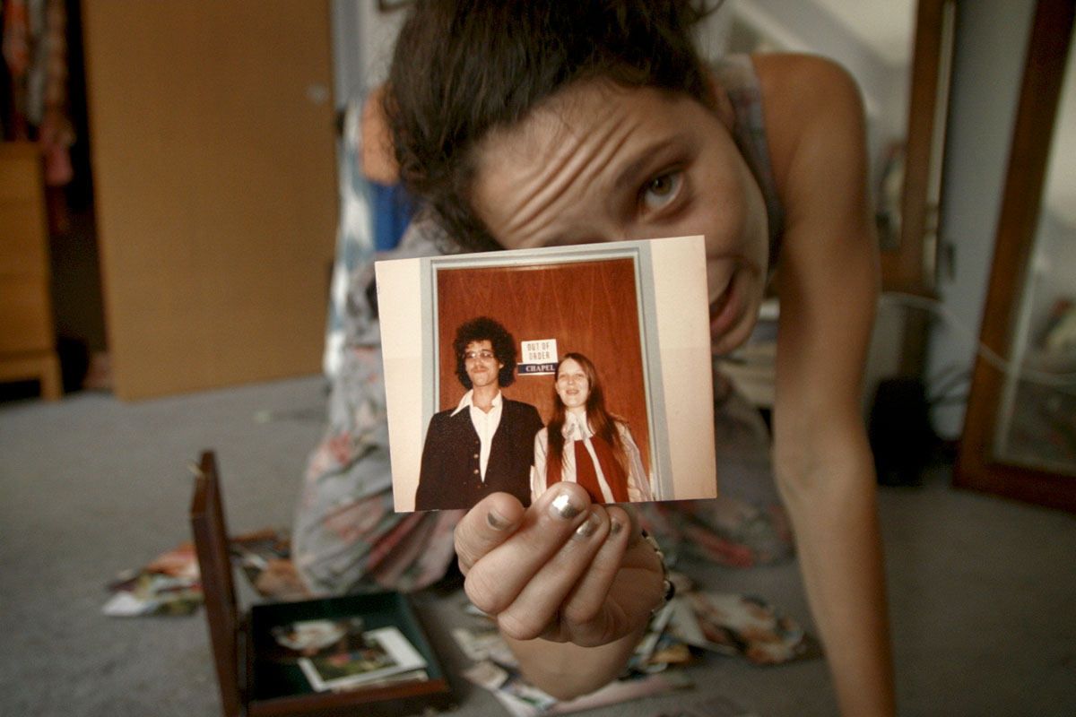 A white woman with dark hair crouched over holding up a polaroid of a man and a woman. From Nira Burstein's 'Charm Circle'. Courtesy of Big Sky Documentary Film Festival. 