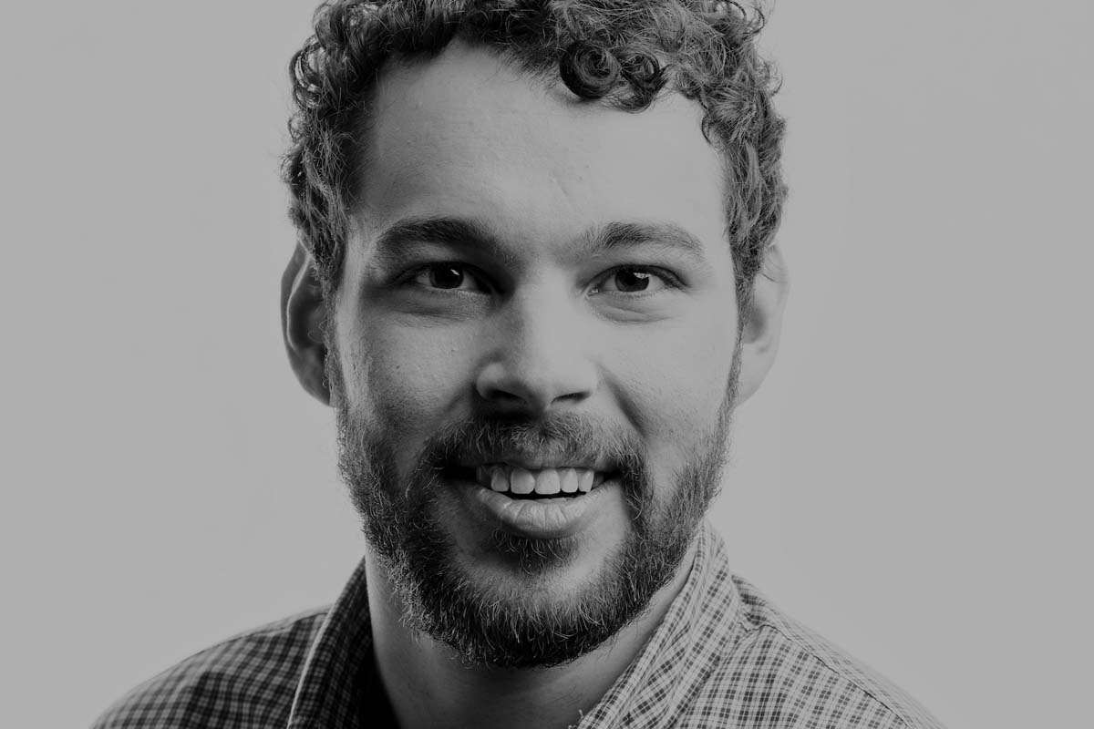 A black-and-white headshot of filmmaker Reid Davenport, a white man with a beard and curly hair, smiling subtly. Photo by Bret Hartman.