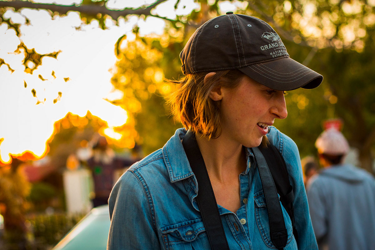 Headshot of a woman in her late 20s wearing a baseball cap over short brown hair. She has a camera hanging around her neck and it is golden hour behind her through the trees. 