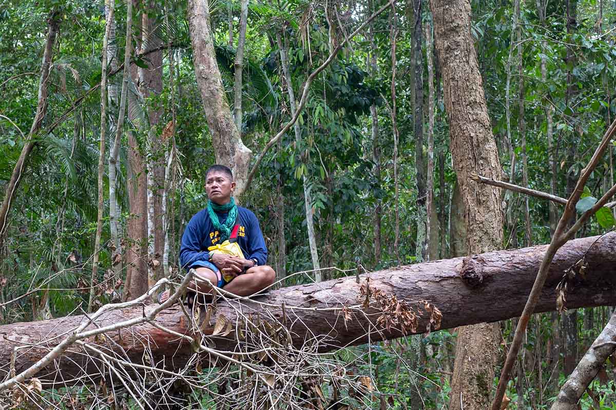Tata is a middle-aged Filipino male activist; he sits on a fallen tree trunk; he is wearing shorts and a blue t-shirt that says "Palawan para-EnviFORCER."  Photo from Karl Malakunas' 'Delikado.' Courtesy of Hot Docs.