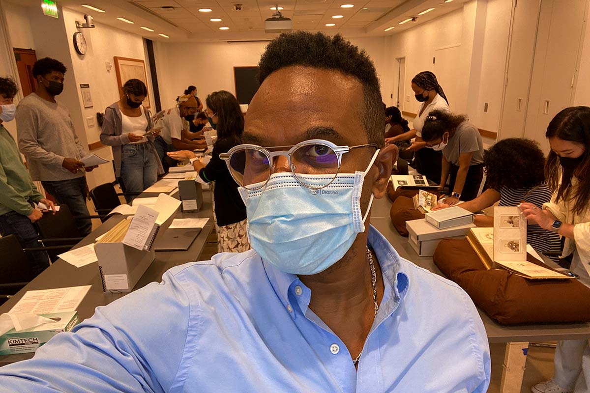Thomas Allen Harris is a Black male filmmaker, seen here wearing a blue shirt, glasses, and a surgical mask. He is taking a selfie, and his students are visible in the background. Courtesy of Thomas Allen Harris.