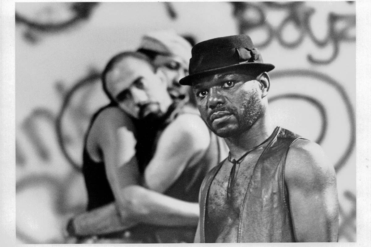 Marlon Riggs is a Black male filmmaker seen here in an archival black and white image from his film 'Tongues Untied.' He is wearing a leather vest and a hat. Courtesy of the Criterion Collection.