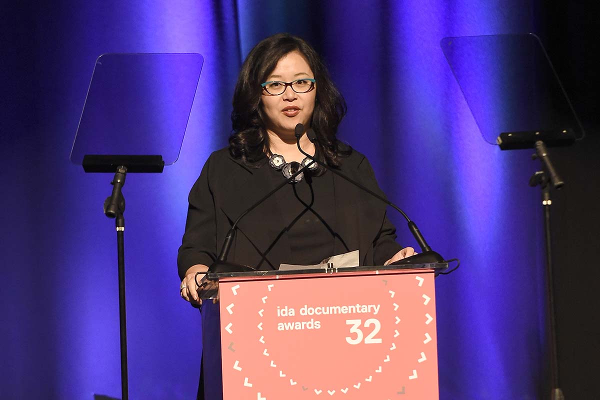 MacArthur Foundation's Kathy Im is an Asian American woman with black hair and glasses, wearing a black dress. She is at the podium at the IDA Documentary Awards. :Photo courtesy of Getty Images. 
