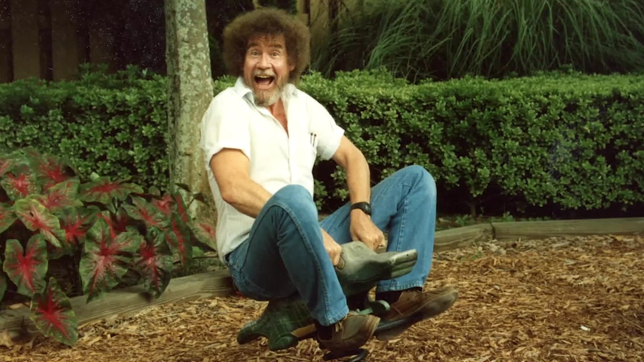 Bob Ross is white artist with a brown afro and a mostly grey beard. He is riding a bouncy alligator ride in a playground. He is wearing a white shirt and blue jeans. Image from Joshua Rofé’s ‘Bob Ross: Happy Accidents, Betrayal & Greed.’ Courtesy of Netflix. 
