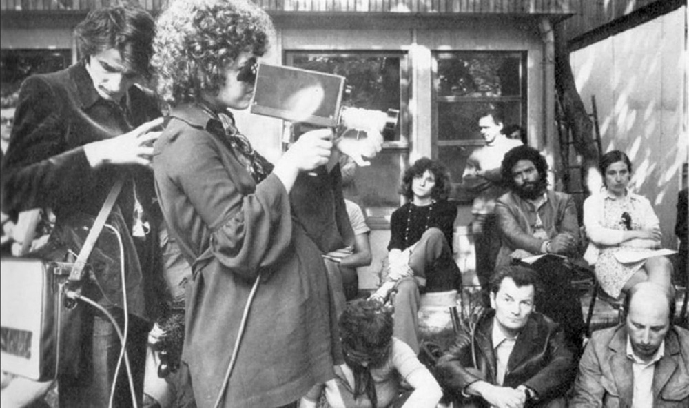 Black-and-white image of French feminist filmmaker Carole Roussoupolos, whose films are now streaming on “Another Screen.” In the photo, she is pregnant and is filming using a handheld camera, with people looking on. Courtesy of Another Screen; Centre Audiovisuel Simone de Beauvoir, Paris.