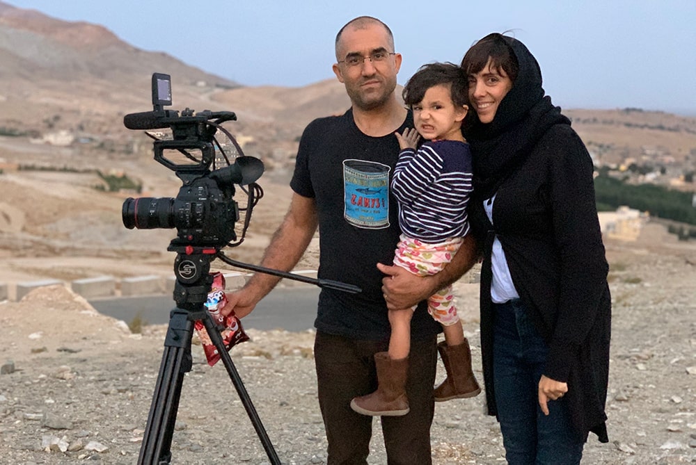 Elizabeth Mirzaei and Gulistan Mirzaei standing in an Afghanistan desert with a camera, in 2019. Gulistan is carrying their daughter, Maryam. Elizabeth is wearing a headscarf, Gulistan is wearing a black t shirt and glasses, Maryam is wearing a striped shirt and floral pants. Image courtesy of the authors.