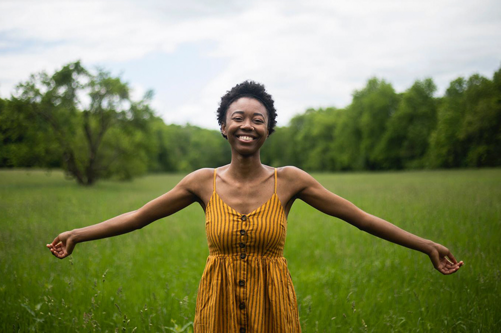 Elodie Edjang, a Black woman with short hair and a yellow dress stands in a grass field.