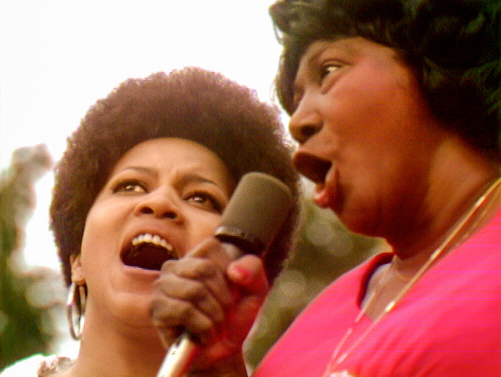 Two Black women singing a duet at the Harlem Cultural Festival. Mavis Staples (left) is wearing hoop earrings and Mahalia Jackson (right) is in a pink shirt. Courtesy of Searchlight Pictures.