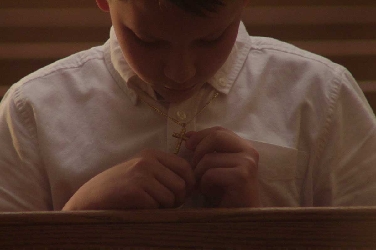 Terrick Trobough is a young white male actor wearing a white shirt, sitting on a church bench. He is wearing a cross. Image from Robert Greene’s ‘Procession.’ Courtesy of Netflix.