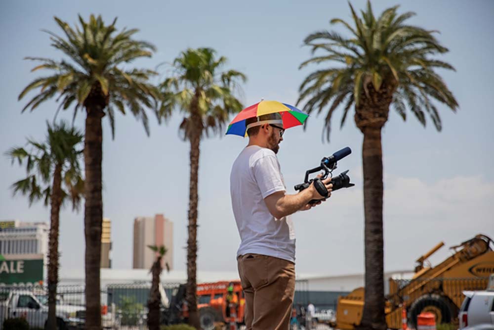 John Wilson is a white male filmmaker wearing a colorful sun umbrella around his head. He is holding a video camera with palm trees in the background. From ‘How To with John Wilson.’ Image courtesy of Joe Buglewicz/HBO.