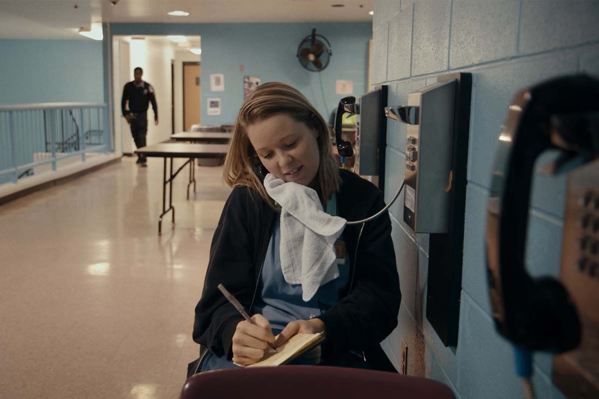 A white woman with shoulder-length red hair talks on a pay phone in a hallway of a correctional facility. From Jennifer Redfearn and Tim Metzger’s ‘Apart,’ which airs February 21 on ‘Independent Lens.’ Courtesy of the filmmakers.