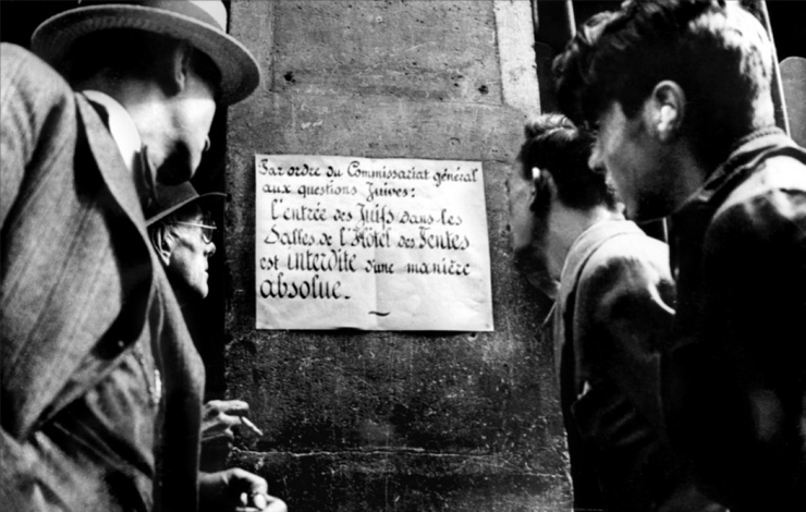 The back of four men in hats staring at a sign posted on the wall. From Marcel Ophuls' Hotel Terminus (1988).