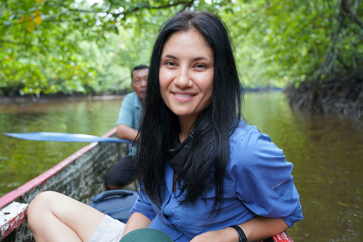 Allison Hanes, a woman with light skin and long black hair, sits in a small boat on a river. An older man with brown skin and dark hair sits behind her. Image courtesy of Hanes.