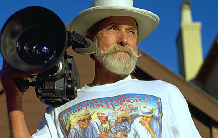 A white man with a beard and mustache wears a white hat and holds his camera on his shoulder.