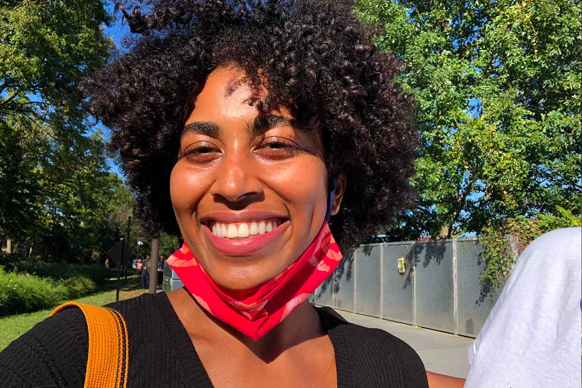 Cydney Tucker is a Black woman with a huge smile and short curly hair. She is seen wearing a red mask on half of her face in a green and luscious park.