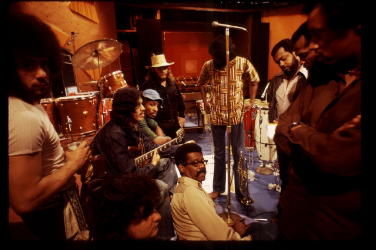 Ellis Haizlip, sits in the middle of the set of his variety show 'SOUL' surrounded by members of the '70s funk band Mandrill, some members of whom are holding their instruments.