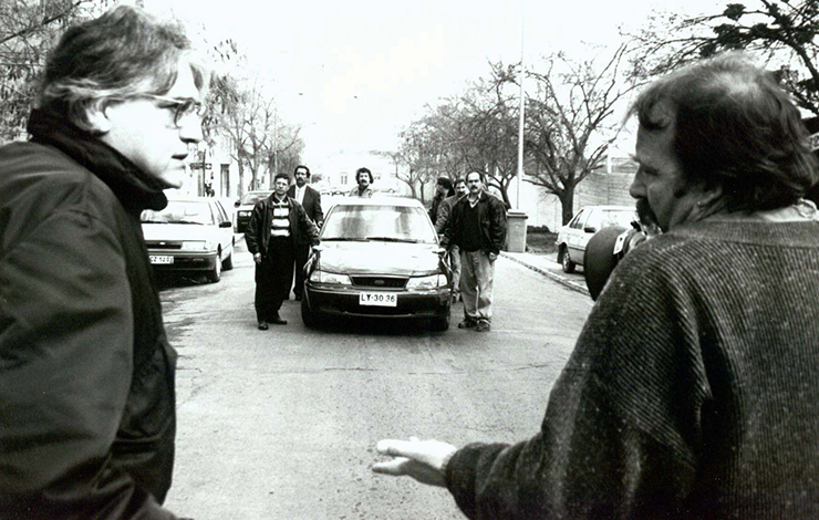 two middle-aged men are talking to each other in the middle of the street, facing a group of men surrounding a car