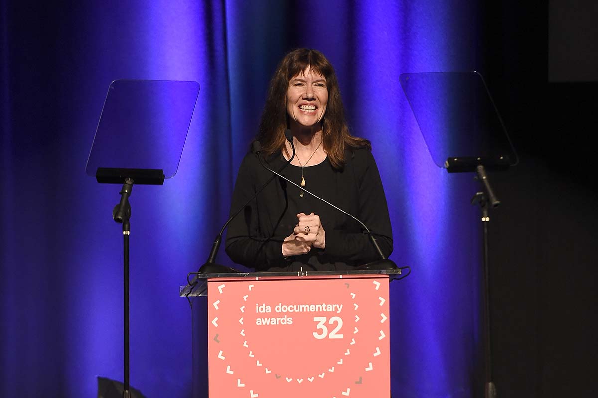 Diane Weyermann speaking at a podium at the 2016 IDA  Documentary Awards. She is wearing black and has her hair untied.