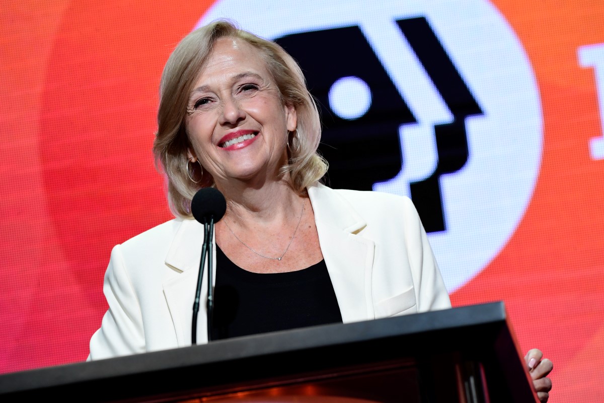 PBS President and CEO Paula Kerger at a podium, wearing a white blazer over a black blouse with the PBS logo behind her.
