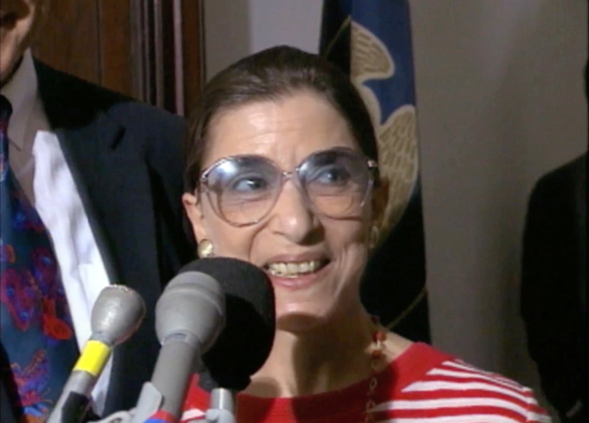 Justice Ginsburg is pictured here at a press conference, in front of a bank of microphones, and she is wearing a red-and-white striped shirt.