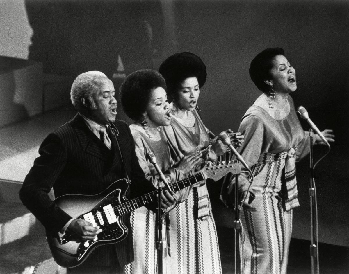 Three Black women in long skirts singing, one Black man playing the guitar. They are on stage. There are three microphones.