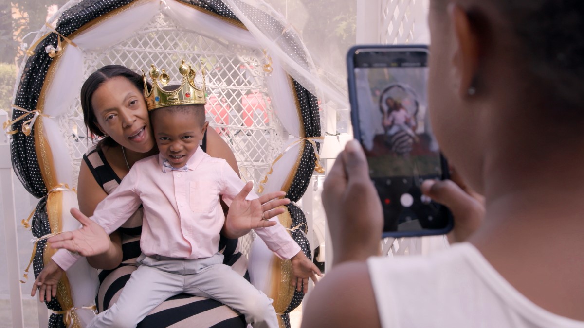 A Black boy, wearing a golden crown and a pink shirt, sits on the lap of a Black woman in a striped dress. A Black girl takes a photo.