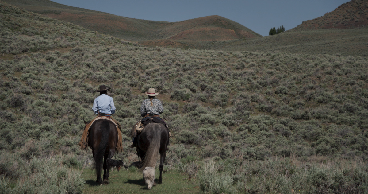 Hollyn Patterson and Colie Moline are two white women wearing cowboy hats and shirts. We can see their backs as they ride horses against the lush greenery of rural Idaho. From Emelie Mahdavian’s ‘Bitterbrush.’ Courtesy of Alejandro Mejia/Magnolia Pictures.