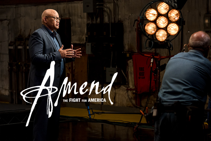 Larry Wilmore, an African American male film director, is wearing a black blazer over a blue shirt, talking to someone off screen on a film set. 