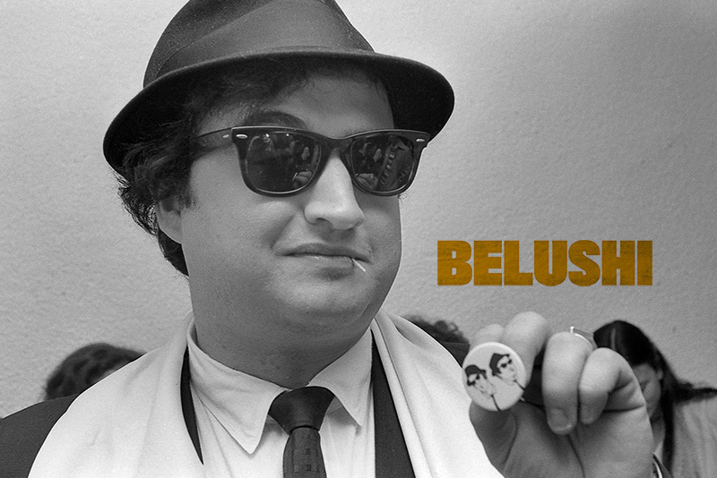 John Belushi is wearing a fedora and black sunglasses, holding up a pin with print of himself