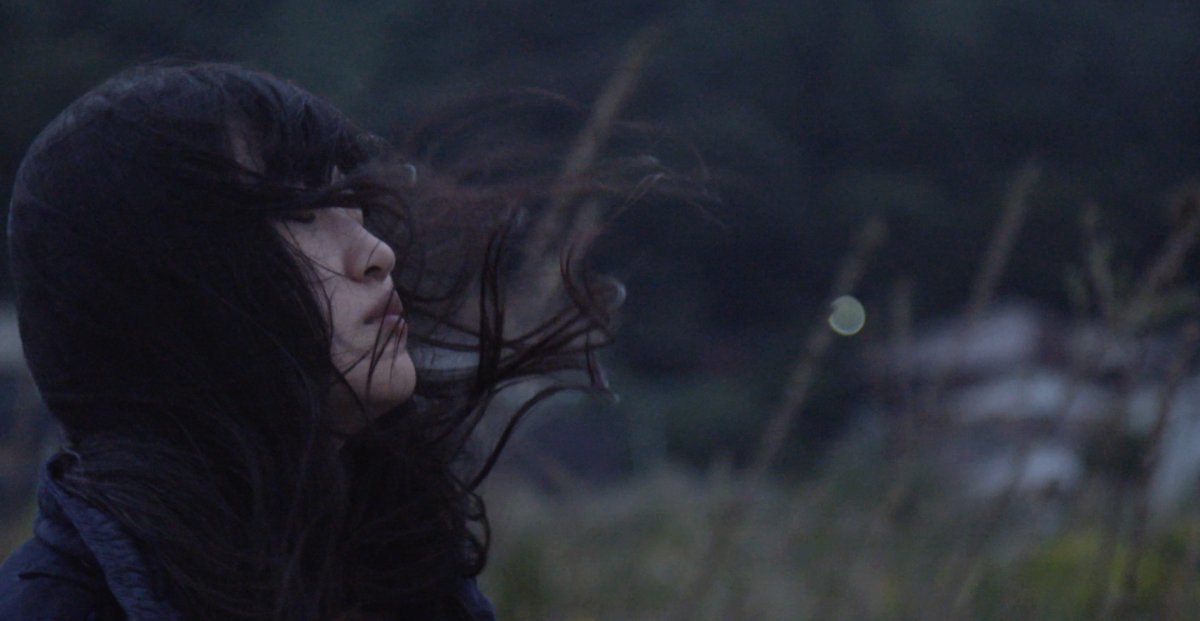 A young Japanese woman stands outdoors with her eyes shut. Her black hair is seen blowing in the wind. From Jennifer Rainsford's 'All of Our Heartbeats Are Connected Through Exploding Stars.' Courtesy of Hot Docs.