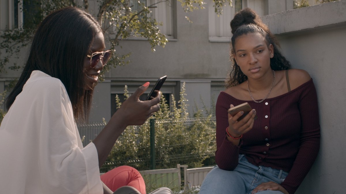 Two young Black women sitting in the sun chatting, and looking at their cellphones. From Alice Diop's 'Nous.' Courtesy of Mubi.