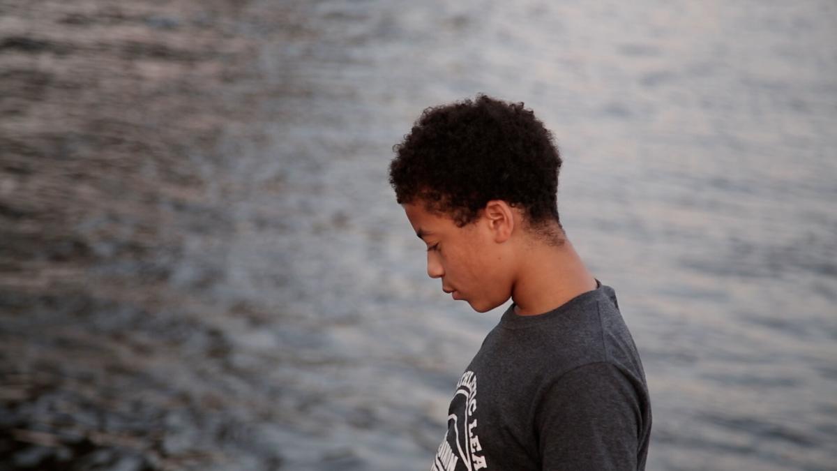 Nyvae Scott, a Black teenager wearing a grey t-shirt, stands by the Schuylkill River in Philadelphia. Image from Lisa Riordan Seville and Zara Katz’s ‘A Woman on the Outside.’ Photo courtesy of the filmmakers  