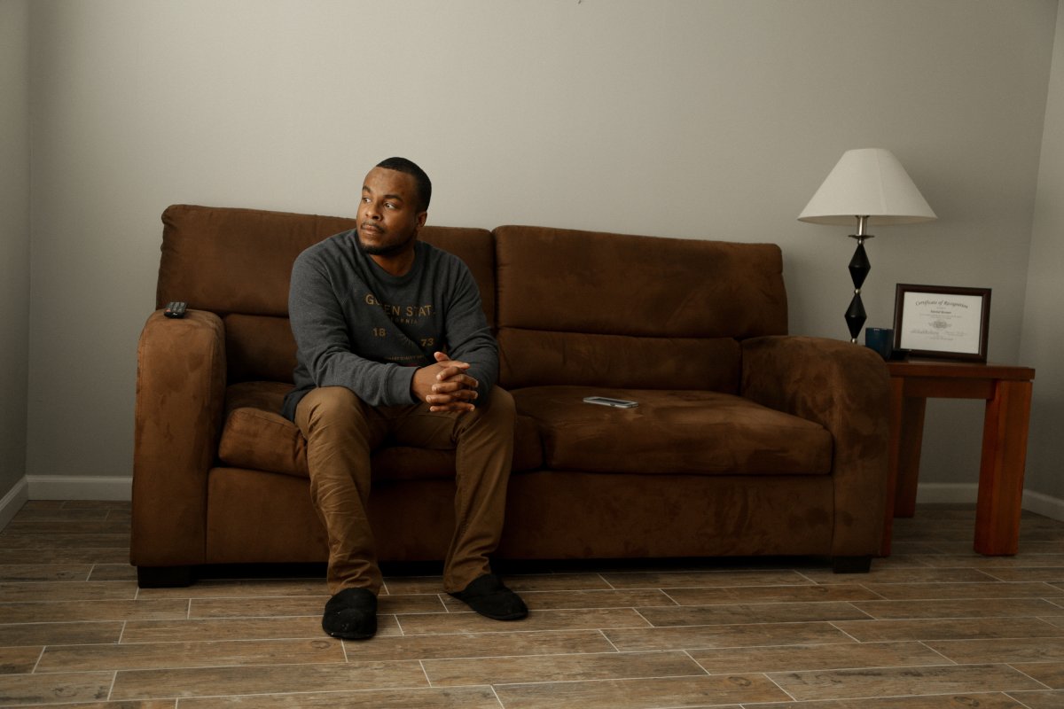 Daniel is a Black male sitting on a brown couch in his apartment in Cicero, IL. He is wearing a gray sweatshirt and brown pants. From Margaret Byrne’s ‘Any Given Day.’ Photo by Margaret Byrne.